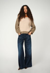 Strickpullover mit beigem Körper und strukturierten braunen Ärmeln, kombiniert mit weit geschnittenen blauen Jeans. Modell mit Händen in den Taschen, neutraler Hintergrund.