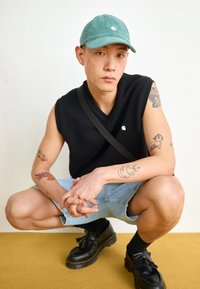 Schwarzer ärmelloser Pullover, hellblaue Jeansshorts, schwarze Schuhe mit dicker Sohle und eine grüne Cordmütze. Sichtbare Tattoos an den Armen.