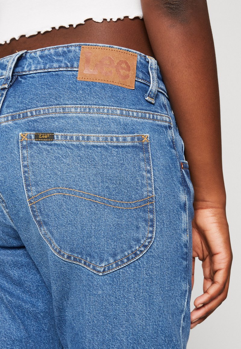 Rückansicht einer Person, die hochgeschnittene blaue Lee-Jeans und ein weißes cropped Top trägt, mit entspanntem Hand an der Seite vor einem hellen Hintergrund.