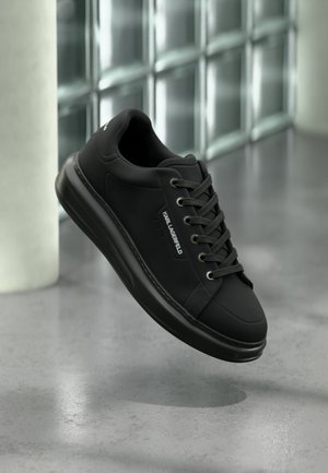 Pantof sneaker negru Karl Lagerfeld cu șireturi negre, talpă de cauciuc și detaliu cu logo, plutind deasupra unui podea gri de beton lângă blocuri de sticlă.