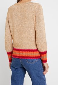 Stickad tröja i beige med en texturerad finish. Har en randig nederkant i röd, orange och beige. Långa ärmar och rund halsringning.