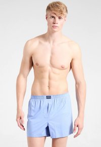 Svetlomodri bombažni bokserji z elastičnim pasom in enim sprednjim odprtjem. Prikazani model je brez majice in pozira nevtralno.