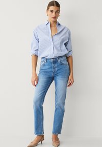 Camisa de botones a rayas azul y blanca con mangas enrolladas, combinada con jeans de mezclilla azul claro. Los jeans tienen un corte recto y un lavado claro.