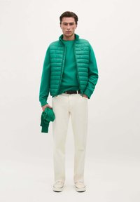 Chaleco acolchado verde sobre un sudadera a juego, combinado con pantalones color crema y zapatillas ligeras. Sudadera verde sostenida en la mano.