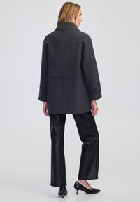 Cappotto grigio in misto lana con una silhouette strutturata, grande colletto e polsini con bottoni. Abbinato a pantaloni in pelle nera e tacchi.