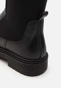 Botín negro con tacón de cuero suave, parte superior de textil blando, suela texturizada y una pestaña vertical para ponerlo y quitarlo con facilidad.