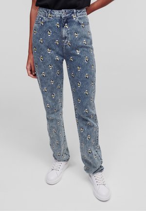 Hellblaue Jeans aus Denim mit kleinen, wiederholten gestickten Pinguinmustern, kombiniert mit weißen Sneakern und einem schwarzen Oberteil.