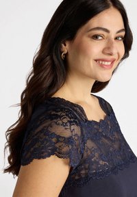 Bluză bleumarin cu detalii din dantelă pe umeri, mâneci scurte și decolteu rotund. Textură de țesătură netedă cu modele de dantelă intricate.