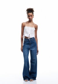 Camisola bianca corta con pizzo, abbinata a jeans in denim blu a vita alta e gamba larga. La modella si trova di fronte a uno sfondo bianco uniforme.