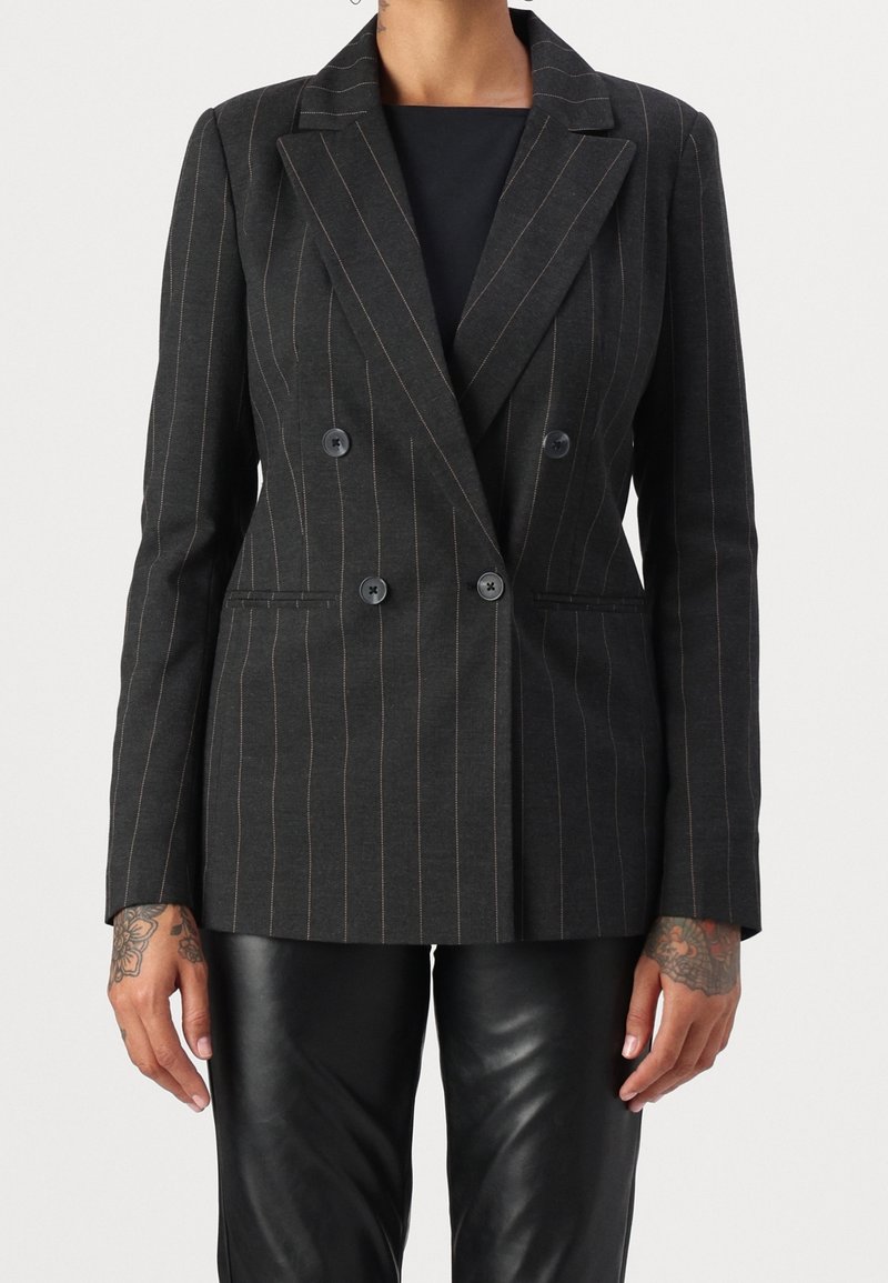 Personne portant un blazer noir à rayures, croisé, une chemise noire et un pantalon en cuir noir, avec des tatouages visibles sur les deux mains.