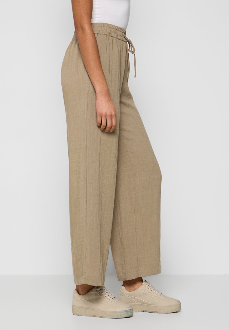 Beige weite Hosen mit elastischem Bund und Kordelzug. Aus einem strukturierten Stoff gefertigt, mit vertikalen Streifen. Kombiniert mit hellen Sneakern.