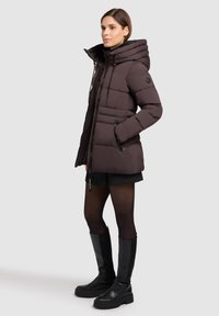 Brun quilted jakke med høj hætte, kontrasterende snore og sidelommer. Båret med en sort nederdel og knælange sorte støvler.