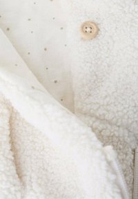Chaqueta de forro polar en blanco roto, que presenta un exterior texturizado, un forro interior suave con patrones de estrellas doradas y un detalle de botón de madera.