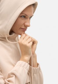 Sudadera con capucha de color beige claro, con una textura suave, que presenta un cierre con cordón y puños elásticos, llevada por un modelo que agarra el cordón.