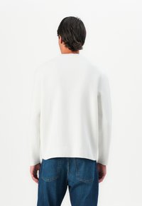 Hombre de espaldas vistiendo una camiseta blanca de manga larga y jeans de mezclilla azul, con un fondo blanco.