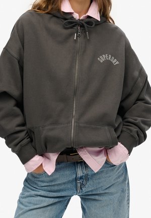 Person wearing a dark gray Superdry zip-up hoodie over a light pink button-up shirt tucked into blue jeans with hands in pockets.