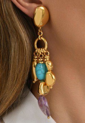 Boucle d'oreille en or avec de grandes pierres turquoise et violettes, portée sur un gros plan de l'oreille d'une personne et ses cheveux châtain clair.