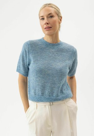 Kvinde med blond hår bundet tilbage iført en blå kortærmet sweater og lyse beige bukser med hænderne i lommerne mod en hvid baggrund.