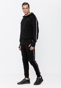 Czarna hoodie na zamek z kangurzą kieszenią i logo na lewej piersi, w połączeniu z czarnymi dresami z paskami po bokach.