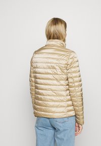 Beige prošívaná bunda s lesklým povrchem, horizontálním prošíváním, stojáčkovým límcem a jemnými švy, nošená s světle modrými džíny.