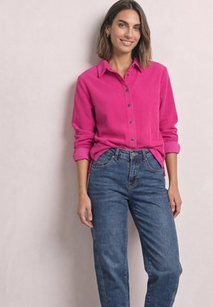 Kvinde smilende iført en lyserød kordfløjls skjorte med knapper og opkrogede ærmer samt blå højtaljede jeans mod en lys baggrund.