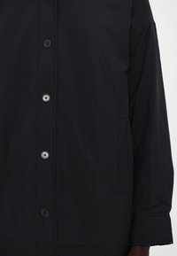 Chemise noire à manches longues avec quatre boutons visibles à l'avant et une poche sur le côté droit.