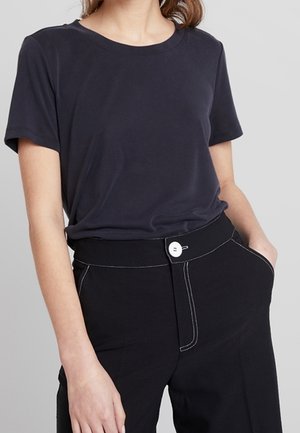 Femme portant un t-shirt bleu marine uni rentré dans un pantalon noir taille haute avec un bouton blanc et des surpiqûres contrastées.