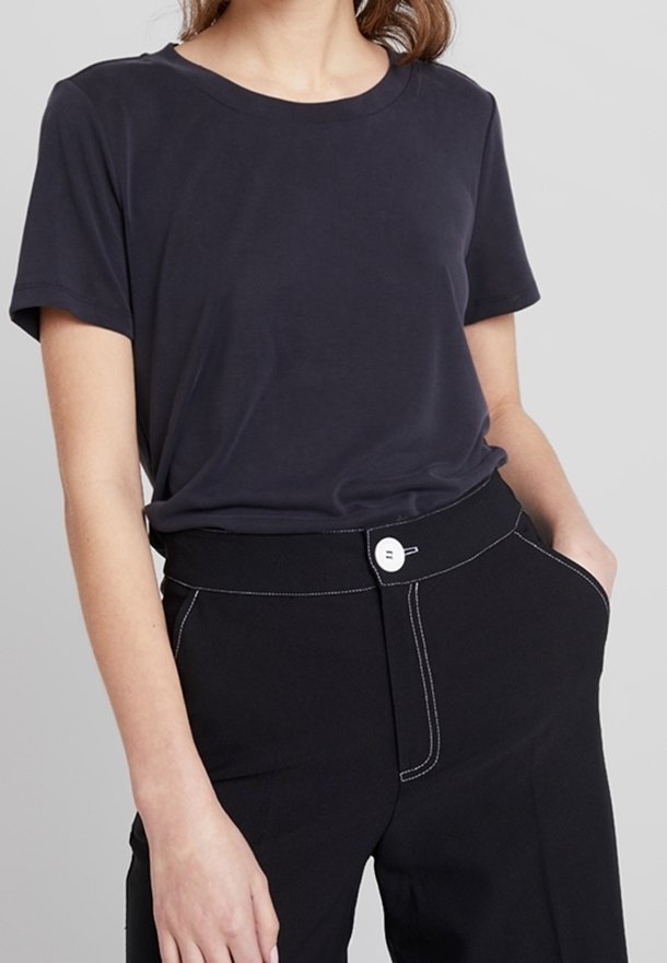 Femme portant un t-shirt bleu marine uni rentré dans un pantalon noir taille haute avec un bouton blanc et des surpiqûres contrastées.
