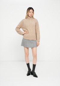 Beige kötött pulóver magas nyakkal, bordás textúrával. Szürke gyapjú szoknyával, sötét zoknikkal és fekete cipőkkel párosítva. A modell egyenesen áll.