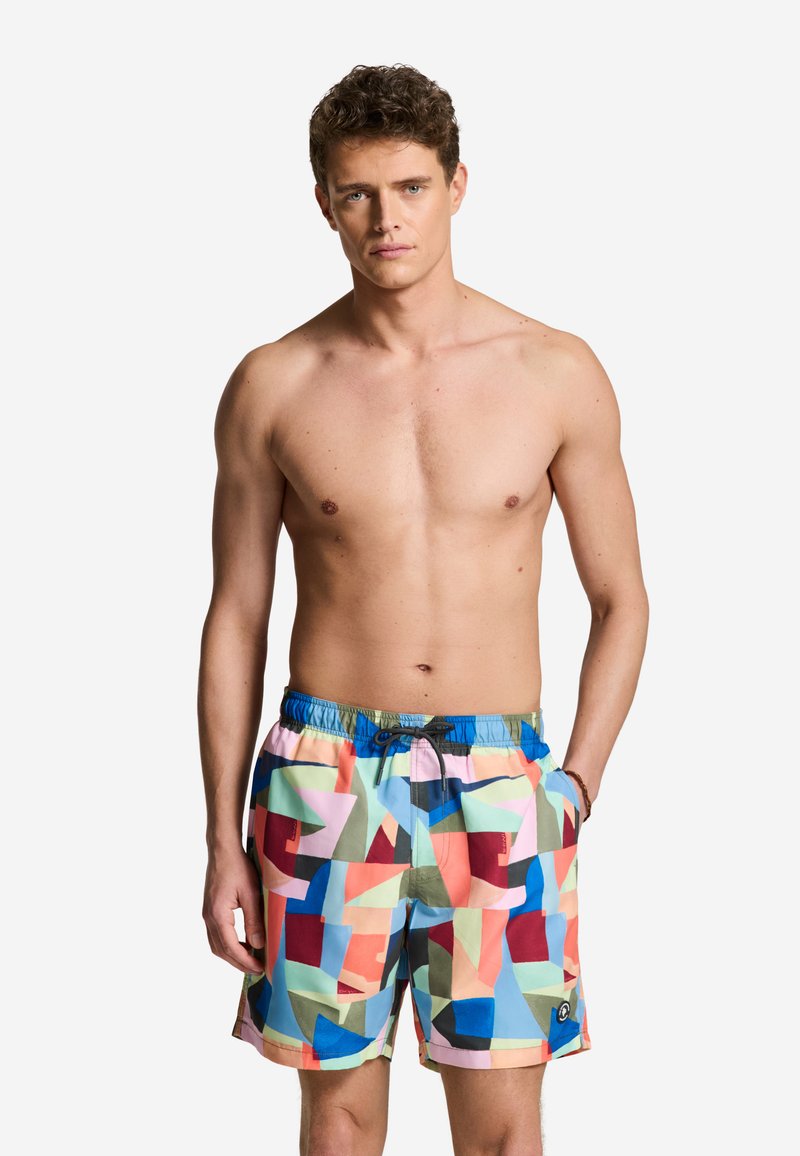 Mann mit lockigem Haar, der bunte Badehosen mit geometrischem Muster trägt, steht mit einer Hand in der Hosentasche vor weißem Hintergrund.