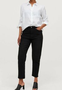 Jeans neri a vita alta con gamba dritta, abbinati a una camicia bianca in lino con bottoni. Le mule nere con tacco completano l'outfit.