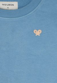 Suéter de punto azul claro con un pequeño detalle de lazo bordado en crema cerca del cuello y una etiqueta blanca de la marca Silbon en el interior del escote.