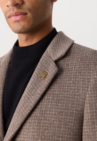 Blazer de lana de cuadros marrones con un cuello con muesca, que presenta un pin de mono en tono dorado en la solapa y un cuello de tortuga negro acanalado debajo.