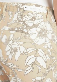 Helle beigefarbene Hose mit weißem Blumenmuster und metallischen Akzenten. Sie verfügt über ein elegantes Design, mit einem Logo am Taillenbund und Taschen auf der Rückseite.