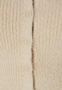 Ljus beige texturerad cardigan med ribbat mönster, fem knappar och mjukt, fluffigt tyg. Närbild av knappslån och texturdetaljer.