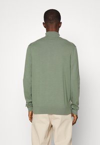Man in een lichtgroene lange mouw turtleneck-trui en beige broek, staand met zijn rug naar de camera tegen een effen witte achtergrond.