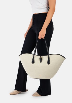 Woman holding large cream tote bag with black handles, wearing black pants, white top, and beige flats, standing on white background.