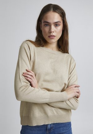 Pull en tricot beige avec un col rond, des poignets en côtes et un ourlet en côtes. Texture douce et coupe décontractée, associé à un jean bleu.