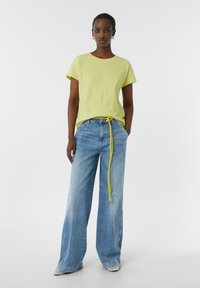 Limonkowa, teksturowana bluzka z krótkim rękawem połączona z szerokimi, jasnoniebieskimi dżinsami, z żółtym paskiem wiązanym i warstwowymi szarymi butami.