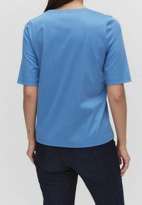 Modré tričko s krátkým rukávem a kulatým výstřihem, hladký povrch a malý ozdobný knoflík na zadní straně. Spárované s tmavými džíny.