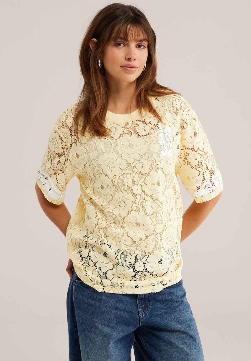 Femme portant un haut jaune en dentelle florale à manches courtes par-dessus une chemise blanche et un jean bleu, debout, les mains derrière le dos.