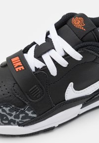 Black sneaker made of leather and mesh, featuring a white Nike swoosh, textured grey accents, and an orange logo near the laces.