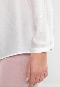Chemise blanche à manches longues avec un tissu texturé, poignet boutonné et ourlet lisse, associée à un pantalon en velours côtelé rose pâle.