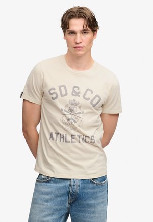 Ung mand med brunt hår iført en lysebeige SD & Co Athletics t-shirt og blå jeans, stående med hænderne bag ryggen.