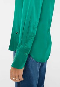Grüne Seidenbluse mit glatter Textur, geknöpften Manschetten und abgerundetem Saum. Verfügt über ein glänzendes Knopfdesign an der Manschette für zusätzliche Gestaltung.