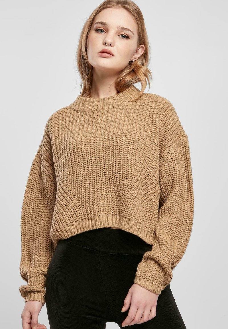 Korte, stevige gebreide trui in beige met een geribbelde structuur, brede mouwen en een ronde halslijn; gecombineerd met zwarte leggings.