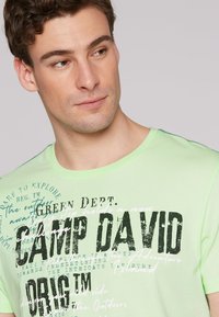 Camiseta de algodón verde claro con texto gráfico en blanco y negro, escote redondo y mangas cortas con una textura sutil.