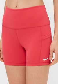 Raudoni sportiniai šortai, pagaminti iš elastingos medžiagos, su aukštu liemeniu, prigludusia siluetu ir maža šonine kišenėle. Matomas baltas Nike logotipas.