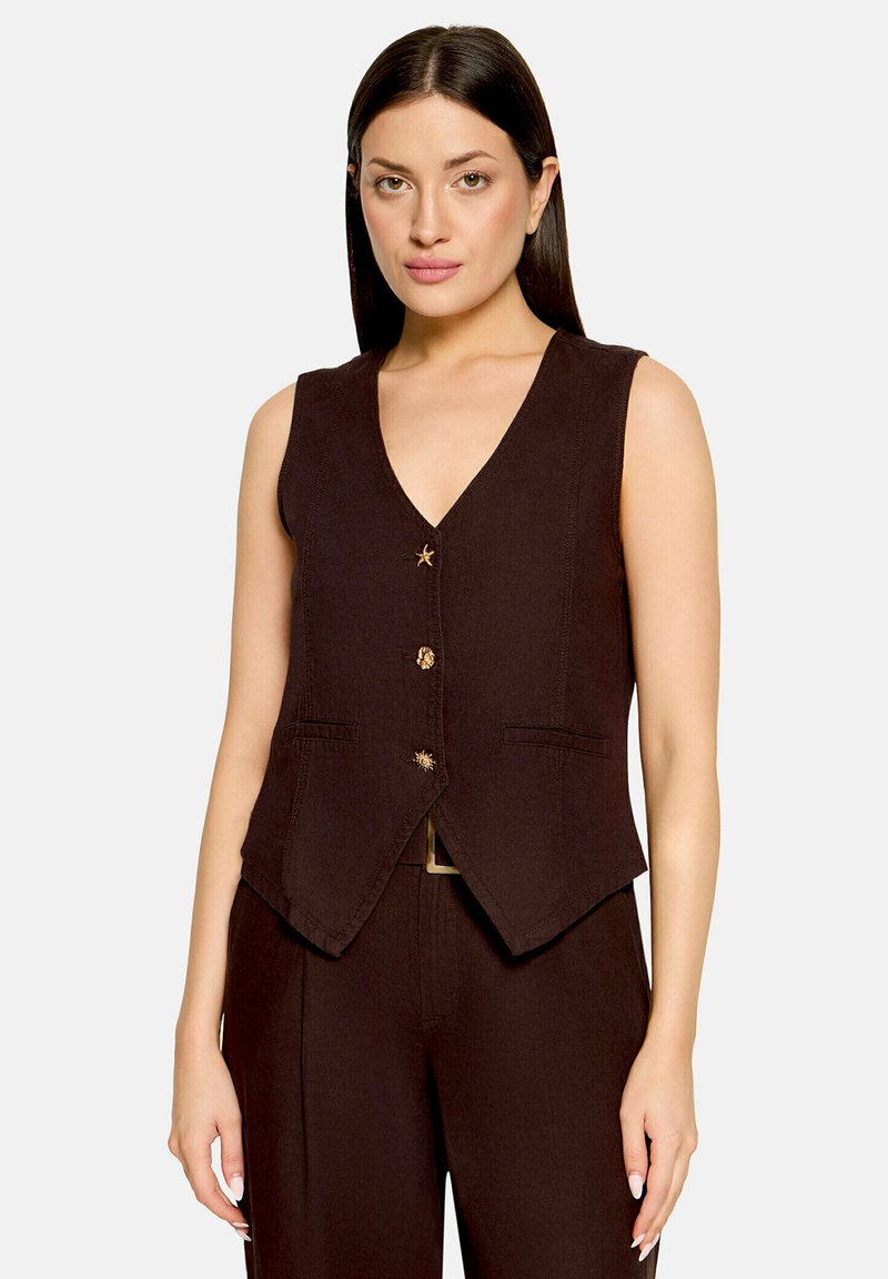 Femme portant un gilet sans manches marron foncé avec des boutons dorés décoratifs sur un pantalon assorti, sur un fond clair uni.