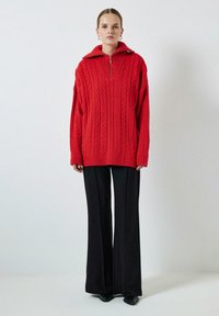 Rode kabelgebreide oversized sweater met een hoge kraag en een halve ritssluiting, gecombineerd met zwarte wijde broek en zwarte schoenen.
